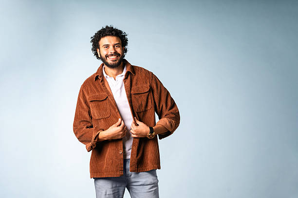 A mid adult Middle Eastern man with a cheerful expression wears a brown jacket and jeans, posed confidently against a soft light blue background.