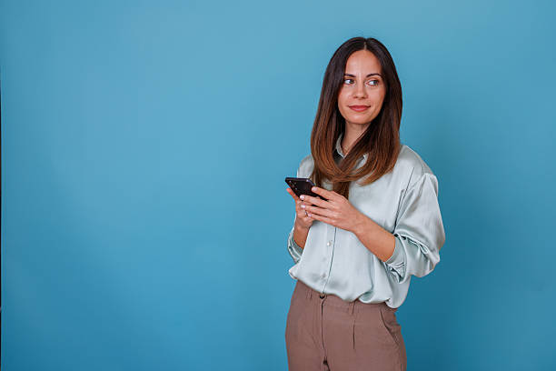 istock Young adult woman using smartphone, looking away casually, standing with copy space on blue background 2247371545