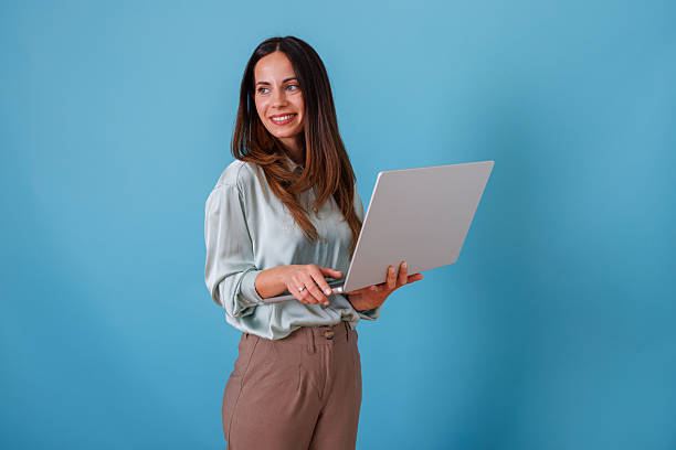 istock Woman standing and holding a laptop, smiling while working remotely. Copy space provided for design on blue background 2247373377