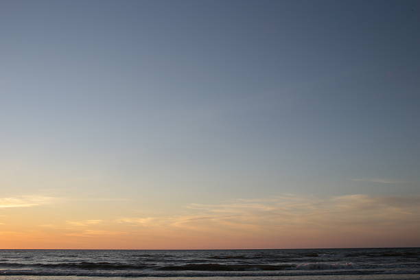 An image focused on the minimalist horizon line where the dark ocean meets the vast sky, displaying a subtle gradient transition from warm orange and yellow near the bottom to cool blue and gray above, perfect for a background.