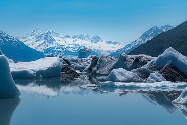 Knik Glacier's icebergs float in the calm water, reflecting the snow-capped mountains under a clear blue sky.
