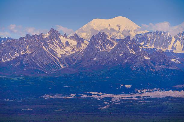 Denali rises majestically above a rugged mountain range under a clear blue sky.