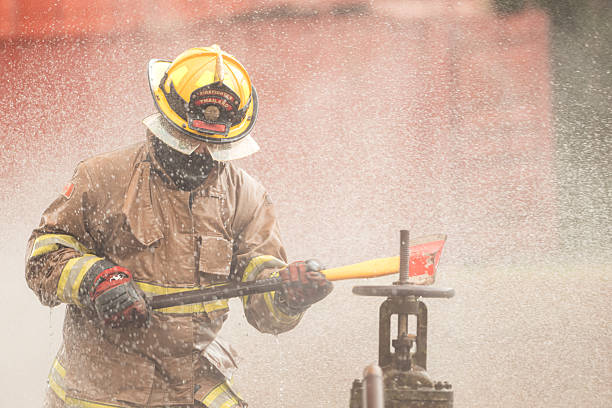 A firefighter in protective gear swings an axe while performing an emergency response in fire hydrant valve. Water sprays through the air as the firefighter works under hazardous conditions