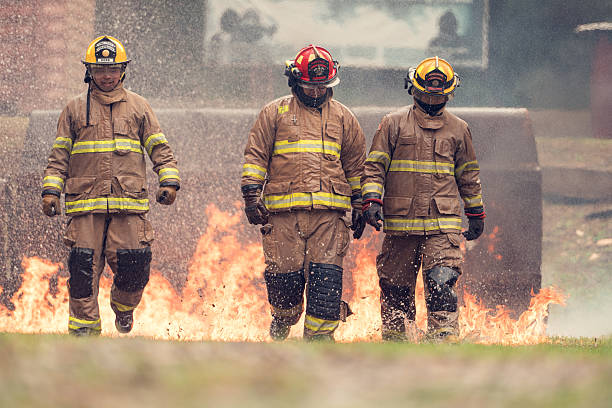 A group of firefighters in full protective gear walks through ground flames during a live fire training drill. Water spray, smoke, and intense heat surround them