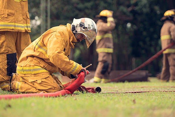 A firefighter in full protective gear kneels on the ground while connecting a fire hose during a training exercise, with other firefighters working in the background