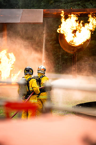 A group of firefighters uses high-pressure hoses to battle a massive blaze during a live fire training operation. Bright flames illuminate the scene as the team works together with focus