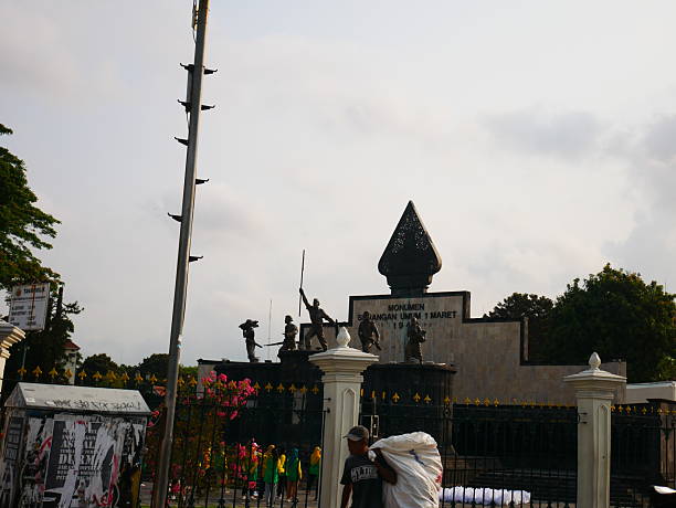 Yogyakarta, Indonesia - October, 2025: the Monumen Serangan Umum 1 Maret, a historical monument located in Yogyakarta, Indonesia, commemorating the 1 March 1949 General Offensive during the Indonesian struggle for independence. The monument features a group of bronze statues representing Indonesian soldiers bravely raising rifles and standing in determined poses, symbolizing the spirit of resistance and patriotism. In the background, trees and a cloudy sky frame the scene, while in the foreground, a man carrying a large sack passes by near the fence adorned with golden spear-tip ornaments. The monument stands as a proud reminder of national heroism and the fight for freedom.
