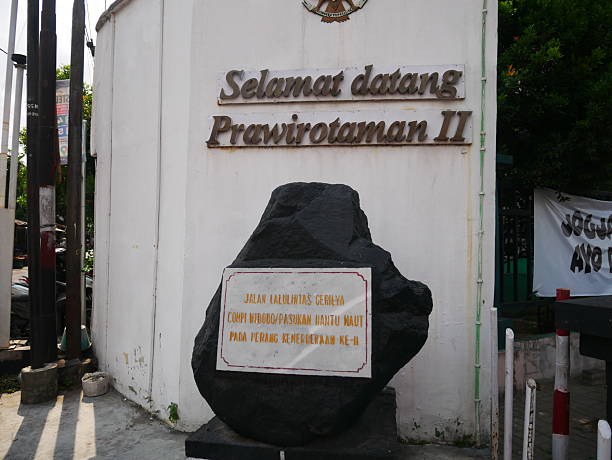 Yogyakarta, Indonesia - October, 2025:  a welcome monument at the entrance of Prawirotaman II in Yogyakarta, Indonesia, featuring a white wall with raised metal lettering reading “Selamat Datang Prawirotaman II.” In the foreground stands a dark stone monument with an inscription commemorating a historical guerrilla route used by Indonesian freedom fighters during the War of Independence. The scene reflects a blend of urban daily life and historical memory, with surrounding street elements such as utility poles, sidewalks, and nearby greenery emphasizing the monument’s role as a local landmark. The image highlights how historical narratives are preserved within contemporary city spaces in Indonesia.