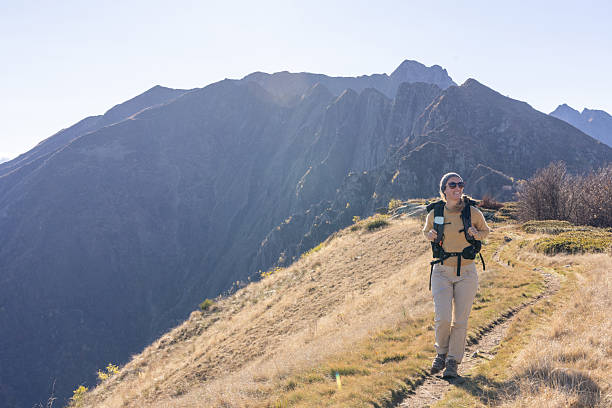 Adult woman hiking on a narrow mountain path with backpack and sunglasses. Solo outdoor adventure in autumn mountain landscape. Active lifestyle and nature exploration.