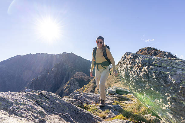 Adult woman hiking on a rocky mountain trail under bright sunlight. Smiling female backpacker wearing sunglasses and outdoor gear. Outdoor adventure and active lifestyle in nature.