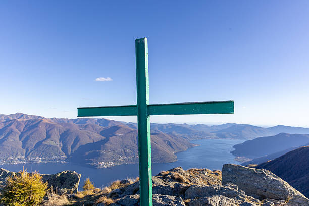 Green metal cross standing on rocky mountain peak with lake and forested hills in background. Scenic nature view with clear sky and distant mountains. Outdoor landscape with spiritual symbol.