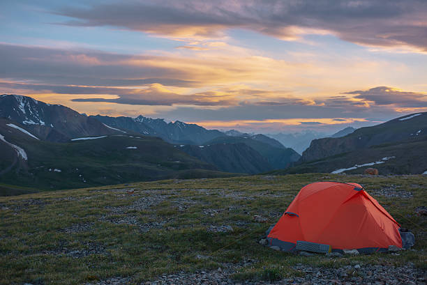 istock Orange tent on grassy stony pass with view to mountain range silhouette far away under clouds in vivid sunset color sky. Scenic alpine landscape with red tent in mountains under colorful cloudy sky. 2247399936