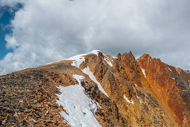 istock Snow-capped mountain ridge with cornice and red sharp rocks on top in sunlight. Snow cap and shiny pointy peak of gold color. Snow dome and colorful rocky peaked top. Sunny cloudy high mountains view. 2247399966