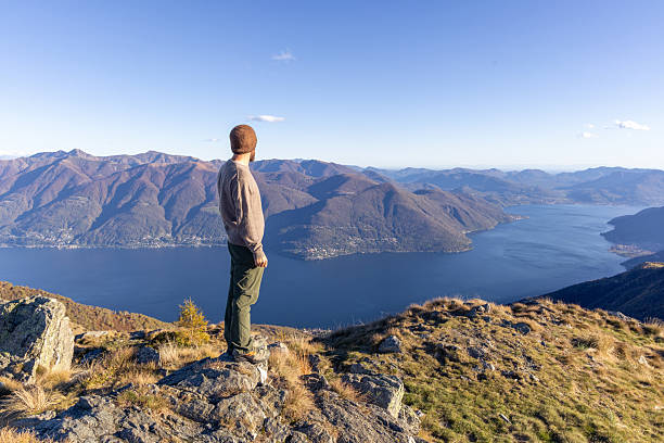 Adult man wearing beanie and sweater stands on rocky mountain peak overlooking a vast lake and mountain range. Scenic outdoor landscape with clear sky and natural wilderness.