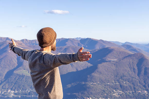 Adult man wearing a brown beanie and sweater stands with arms wide open facing mountain range. Person embracing nature and fresh air in outdoor scenic landscape. Freedom and joy in nature.