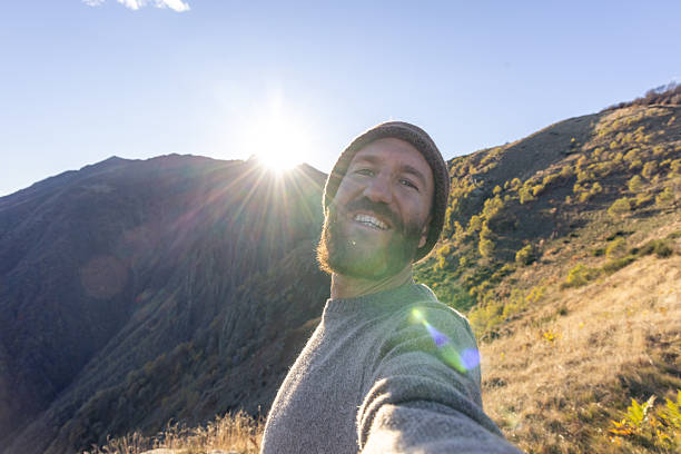 Smiling adult man with beard and beanie taking selfie outdoors on mountain trail. Happy hiker enjoying nature and sunlight in autumn landscape. Outdoor adventure and active lifestyle.