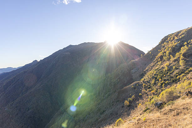 Bright sunlight emerging from behind a mountain ridge in a clear blue sky. Scenic mountain landscape with dry grass and sparse trees on hillside. Nature and outdoor scenery.