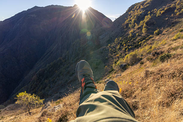 Person wearing hiking shoes resting on grassy mountain slope at sunset. Scenic outdoor moment with sun setting behind rugged peaks. Relaxation and nature appreciation in wilderness.