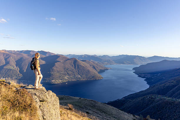 Adult woman standing on rocky cliff with backpack overlooking large lake surrounded by mountains. Solo hiker enjoying nature and scenic outdoor adventure. Mountain landscape with clear sky.