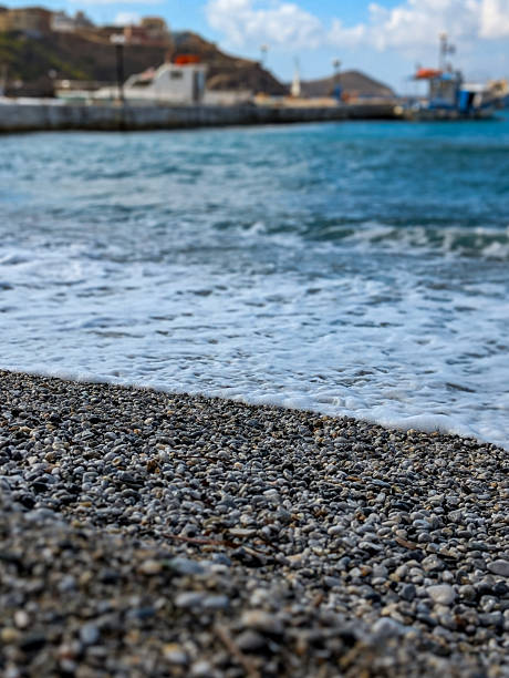Pebble beach shoreline with gentle waves and foamy surf, blurred harbor and seaside town on the horizon under a sunny blue sky