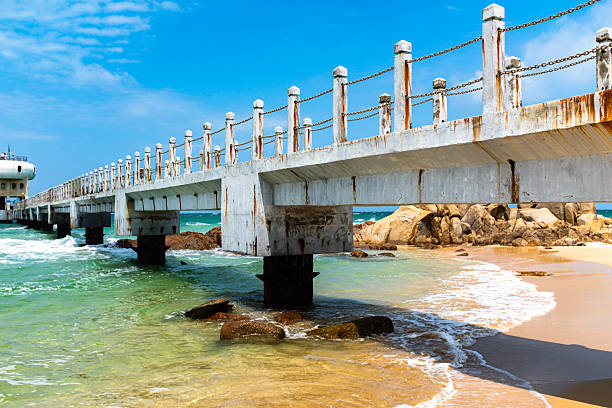 This bright coastal landscape captures a long, straight concrete pier or bridge jutting out into the ocean. The structure features vertical posts connected by simple metal chains and shows visible signs of aging, with rust stains streaking down its white sides. Below, crystal-clear water transitions from the golden hues of the sandy bottom to a vibrant turquoise green as it laps against the shore and large scattered rocks. The scene is set under a vivid blue sky with wispy clouds, creating a sunny and serene seaside atmosphere.