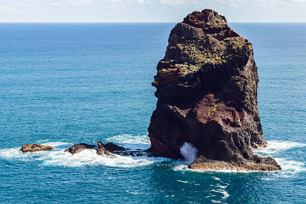 Isolated volcanic sea stack near Sao Lourenco Point in Madeira, rising from deep blue Atlantic waters with waves crashing at its base and sunlight revealing rich textures of lava rock