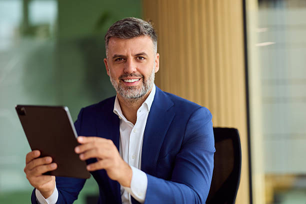 istock Confident Businessman in Blue Suit Smiling While Holding Tablet in Modern Office Environment 2247415320