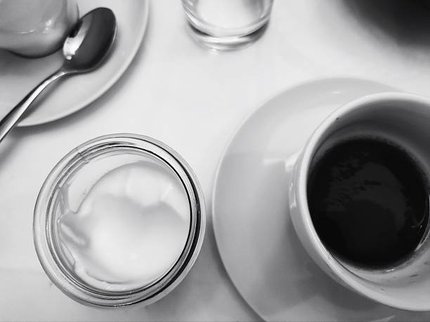 A black and white composition featuring a cup of coffee, a small container of cream, and a teaspoon on a white surface. The overhead view creates a minimalist, geometric layout.