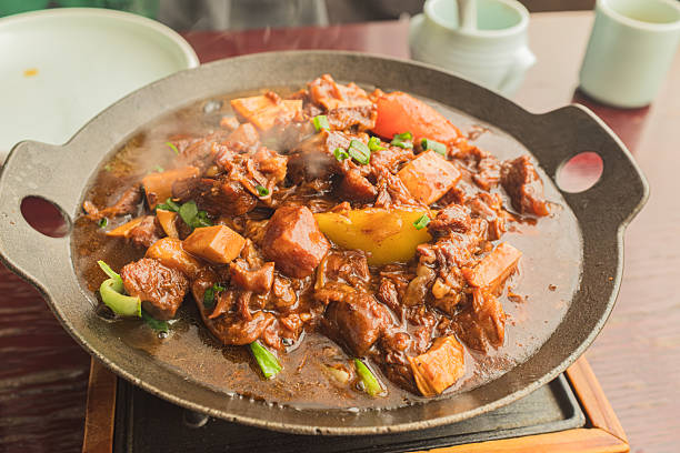 This high-angle photograph captures a rustic and hearty dish of braised beef, served sizzling hot in a shallow black cast-iron pan resting on a wooden base. The stew is composed of tender chunks of dark beef, soft yellow potatoes, and bright orange carrots, all enveloped in a thick, savory brown gravy. Freshly chopped green onions garnish the dish, providing a vibrant color contrast. Visible steam rises from the food, emphasizing its heat and freshness, while the blurred background hints at a casual dining setting with ceramic cups.