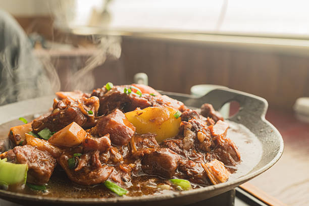This appetizing food photograph highlights a classic comfort dish of braised beef stewed with potatoes, served piping hot in a rustic black cast-iron pan. Wisps of steam rise vigorously from the meal, emphasizing its warmth and freshness. The dish features tender chunks of beef and soft, golden-yellow potatoes coated in a rich, glossy dark brown sauce. A sprinkling of fresh green onions garnishes the top, adding a vibrant color contrast to the savory ingredients, while the bright, blurred background suggests a cozy daytime dining setting.