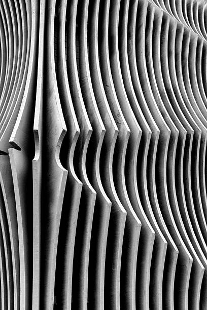 This black and white abstract photograph captures the rhythmic geometry of a modern building facade or architectural sculpture. The composition focuses on a series of repeating vertical fins or blades, likely made of concrete, that curve in and out to create a fluid, wave-like visual effect. The interplay of stark light and deep shadow emphasizes the three-dimensional depth and smooth texture of the material, giving the rigid structure a sense of organic movement.