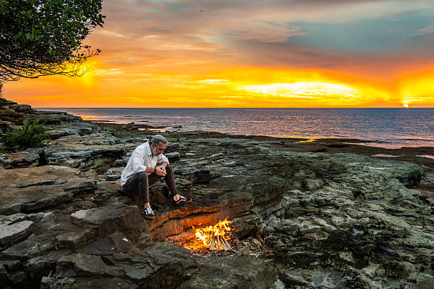 istock Man Sitting by Campfire, Gazing at Sunset over Ocean 2247427300
