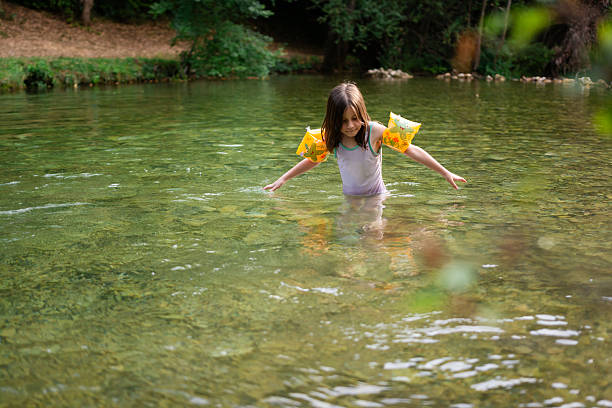 Little girl with armbands standing in clear green river water, smiling and enjoying a carefree summer day outdoors (6–7 yrs)