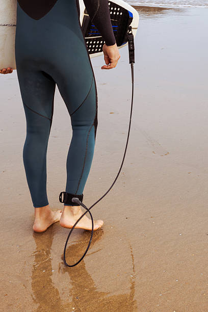 istock Surfer legs in wetsuit standing on wet sand with board leash 2247438125
