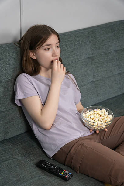istock Young girl sitting on a comfortable sofa, holding a bowl of popcorn while watching a dull movie on tv, feeling tired and alone in her cozy apartment. Vertical photo 2247441263