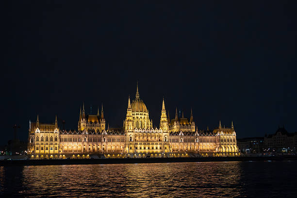 Budapest, Hungary - December 7, 2018: The Hungarian Parliament Building, a stunning Gothic masterpiece on the Danube, shines brightly at night. Its intricate spires and ornate details reflect beautifully in the river, showcasing Budapest's architectural grandeur. This iconic landmark invites admiration, highlighting its historical significance against a picturesque, starry sky backdrop.