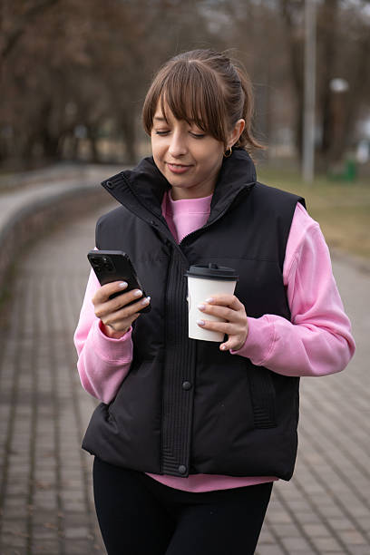 istock Young woman walking through a park, holding a takeaway coffee cup while browsing her smartphone, smiling at something delightful on the screen, enjoying the fresh air and nature. Vertical photo 2247445655