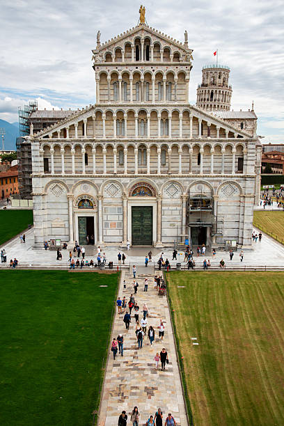 A grand view of Pisa's cathedral facade with the Leaning Tower in the background. Tourists stroll the marble grounds as a stone path leads toward the historic site, showcasing Italian architecture.
