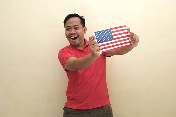 Smiling Southeast Asian man holding US flag, symbolizing immigration, visa, study abroad or citizenship, isolated for promotional or ad use.