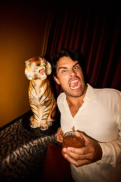 Playful young man holding a drink while looking at camera with an exaggerated expression beside a decorative tiger statue in a warmly lit New Year’s Eve lounge.