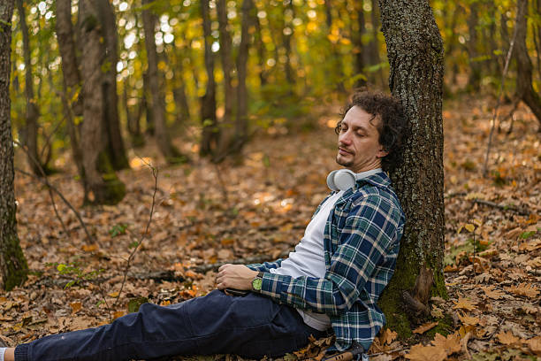 Man with wavy hair and closed eyes resting in nature, leaning on a tree trunk. Vacation and healthy lifestyle concept.