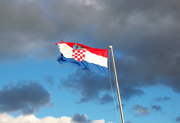 High-resolution image of the Croatian national flag featuring the recognizable red, white, and blue tricolour with the central coat of arms. The flag waves dynamically in the wind, showing slight wear along the edges, adding realism and character. Set against a dramatic cloudy sky, this photo conveys themes of patriotism, national identity, sovereignty, European culture, and travel. Ideal for editorial use, presentations, tourism content, political topics, and cultural representation.