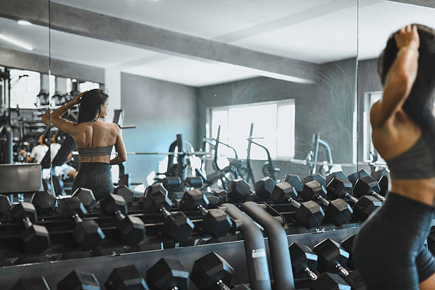 A focused woman in workout attire stands before a gym mirror, surrounded by dumbbells and fitness gear, capturing a moment of strength, dedication, and motivation during a training session.
