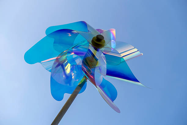 A bright blue wind spinner twirls in the gentle breeze under a clear sky, reflecting sunlight and creating a playful display of colors. The scene captures a cheerful atmosphere.