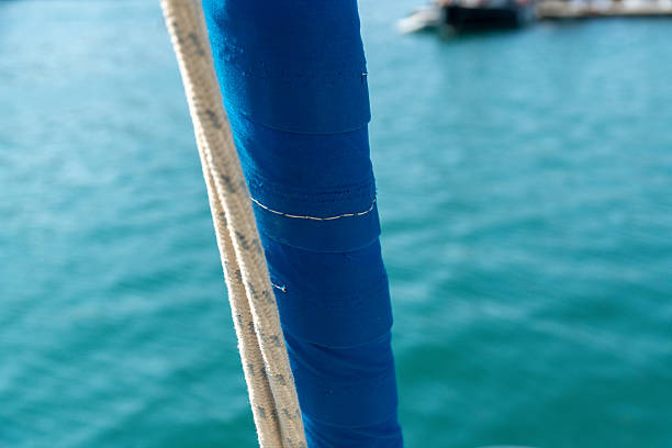 Close-up view of a wrapped blue sail cover with visible stitching next to a rope on a sailboat, against blurred turquoise water background.
