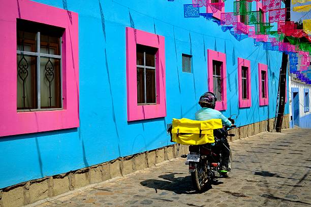 Oaxaca de Juarez, Oaxaca, Mexico – November 7, 2025: A delivery motorcyclist rides past a bright blue building with pink window frames and papel picado above.