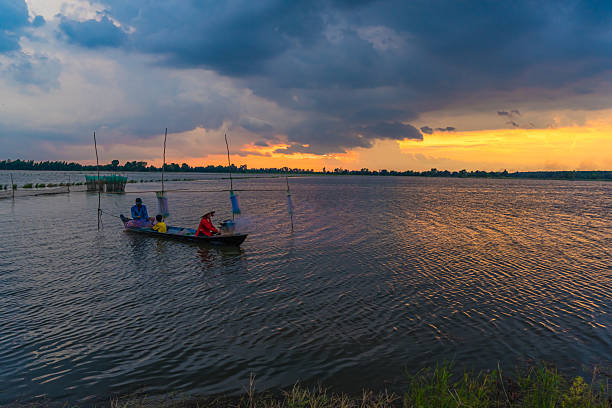 Long An, Vietnam - Aug 22, 2024: A beautiful, serene sunset. A local family a father, a child, and a mother are seen on a small wooden boat, tending to their nets or fishing as the sun dips below the horizon