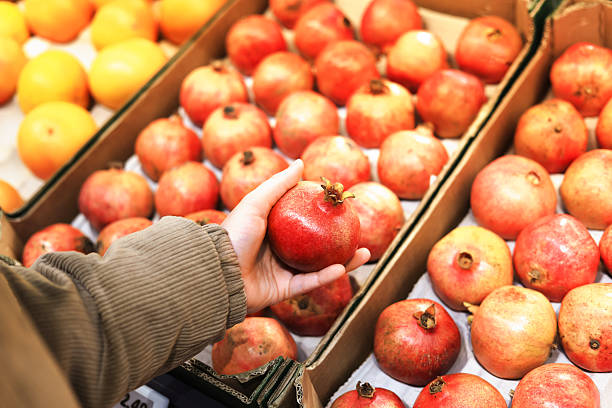 istock A person's hand selects a ripe, red pomegranate from a display box, with blurred oranges visible in the background of the market. 2247488957