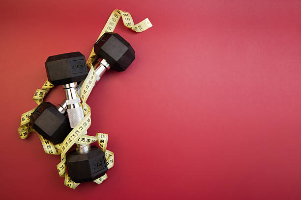 Black dumbbells are placed on a vibrant red background, intertwined with a yellow measuring tape, symbolizing fitness, health, and exercise, perfect for wellness themes