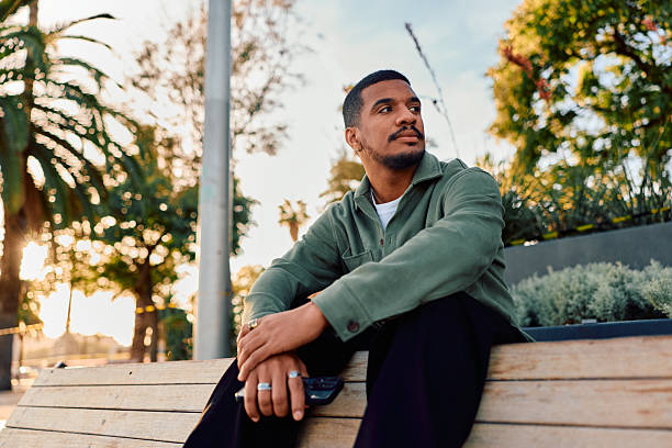 istock Young adult man contemplating while sitting outdoors on park bench 2247492111
